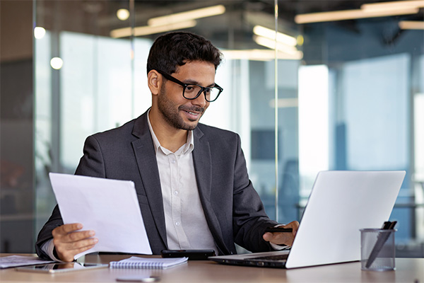 A businessman is reviewing information on his laptop in the office while holding a document.